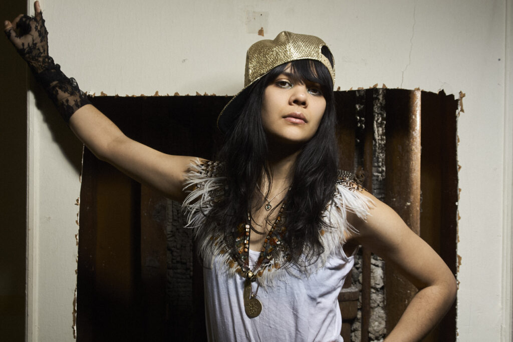 Portrait of musician Bat for Lashes (Natasha Khan) in a gold cap, photographed in New York City by Bert Spangemacher against an industrial backdrop.